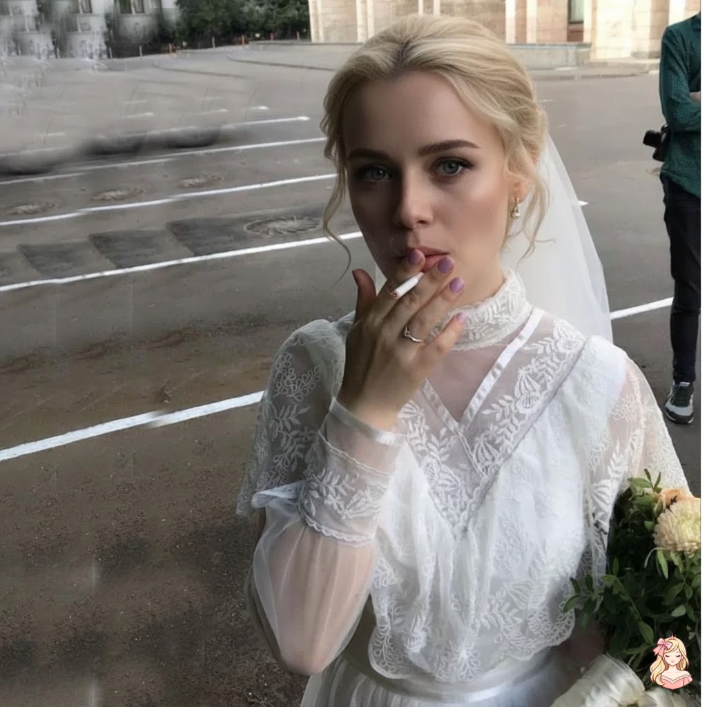 Woman in a white wedding dress with a veil, holding a bouquet and a cigarette, standing on a street.